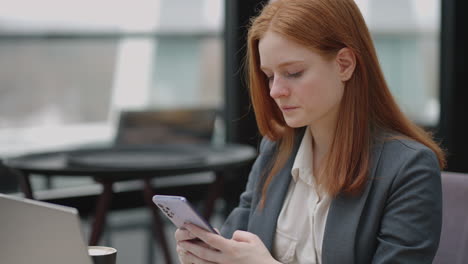 pretty-redhead-woman-in-grey-jacket-is-working-in-co-working-area-viewing-social-media-and-message-in-smartphone